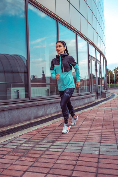 Beautiful young athletic girl in a jacket runs in the morning for a jog, fitness training, sportswear. In the summer in city workout. Background sky and glass windows. - Valokuva, kuva