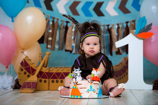 Little cute girl in costume of Apache Indian chief and in a feather headdress sits and tastes a sweet cake. First birthday 1 year and smash cake - Foto, afbeelding