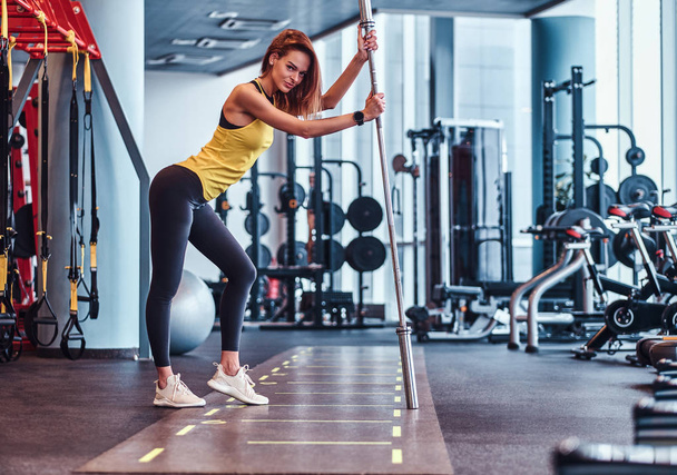 Fitness girl posing for a camera while leaning on a barbell in the modern gym - Photo, Image