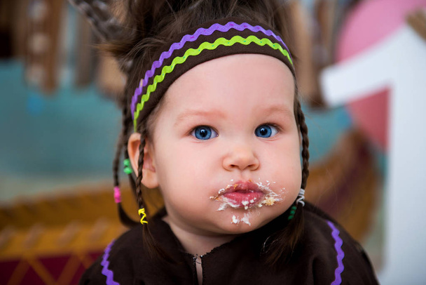Little cute girl in costume of Apache Indian chief and in a feather headdress sits and tastes a sweet cake. First birthday 1 year and smash cake - Φωτογραφία, εικόνα
