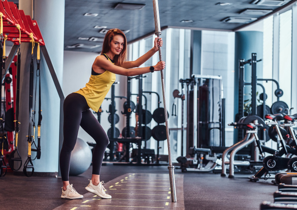 Fitness girl posing for a camera while leaning on a barbell in the modern gym - Valokuva, kuva