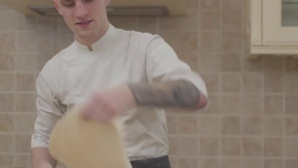 Young guy in chef uniform spinning and tossing disk of pizza dough in kitchen at home. Professional pizzaiolo making pizza. Concept of food preparation - Materiaali, video