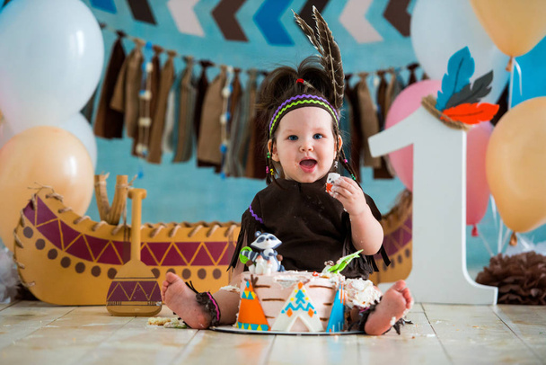 Little cute girl in costume of Apache Indian chief and in a feather headdress sits and tastes a sweet cake. First birthday 1 year and smash cake - Foto, afbeelding