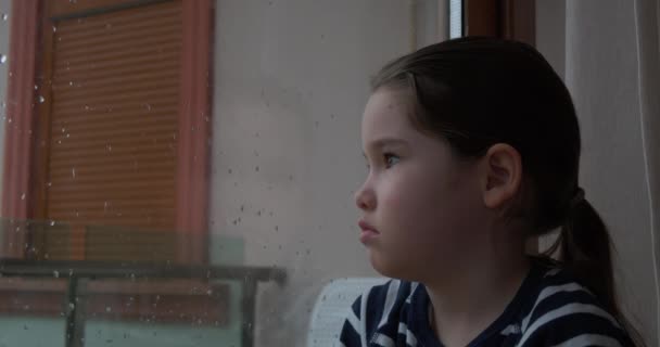 Mała dziewczynka siedzi przy oknie w deszczowy dzień - Materiał filmowy, wideo