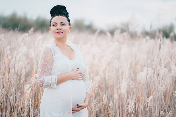 Pregnant beautiful woman in white lace boho dress standing in reeds and holding her belly - Foto, imagen