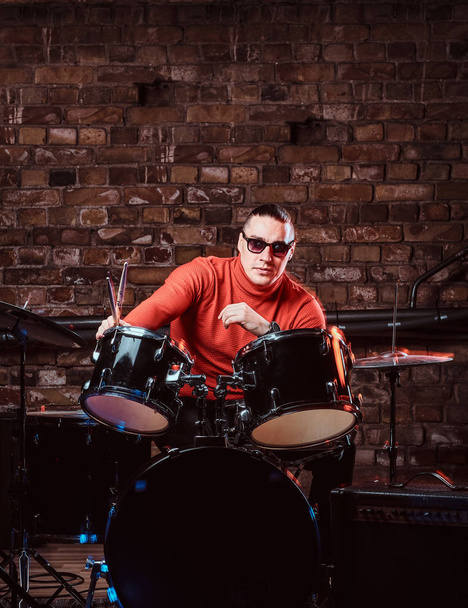 Musicista elegante in occhiali da sole si siede dietro il tamburo contro un muro di mattoni, eseguire in night club
 - Foto, immagini