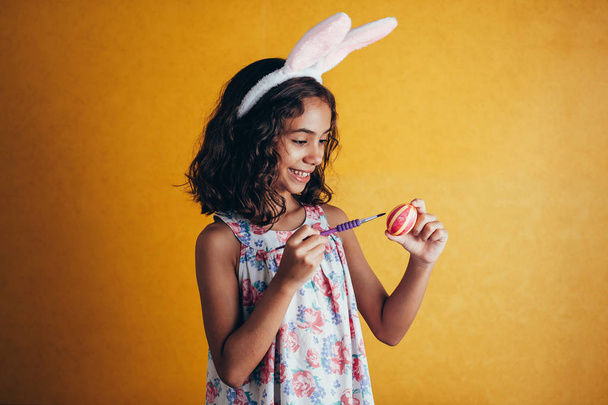 A happy girl painting Easter eggs over color background - 写真・画像