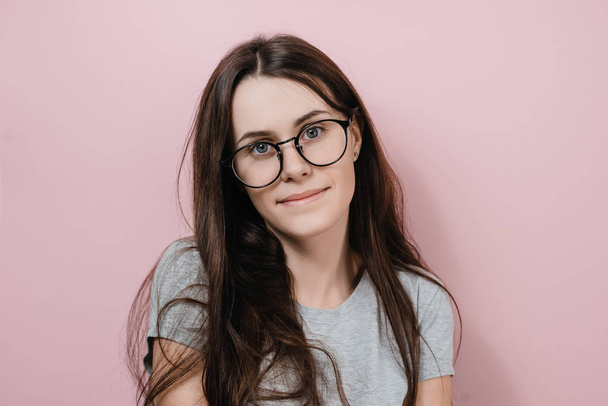 Portrait young attractive girl in glasses feels happy, dressed in gray T-shirt, has long hair, poses against pink wall, looks with mysterious look. People, beauty and skin care concept - Photo, Image