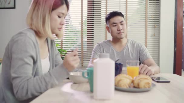 Happy sweet Asian couple man in a wheelchair having breakfast, cereal in milk, bread and using mobile phone reading news after wake up in the morning. Husband and his wife eating food together. - Záběry, video