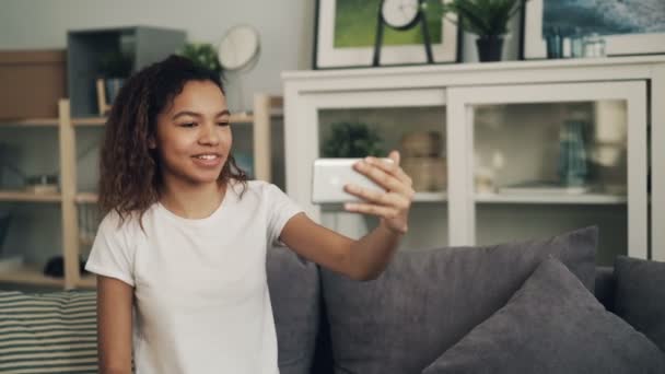 Smiling young African American woman is making online video call looking at smartphone screen talking and laughing sitting on couch in nice apartment. - Imágenes, Vídeo