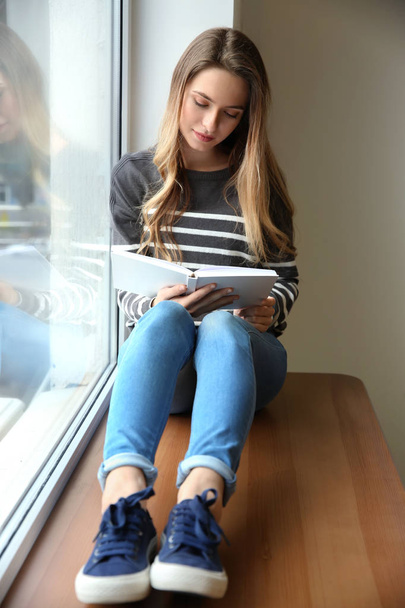 Beautiful young woman reading book while sitting on window sill - Фото, изображение