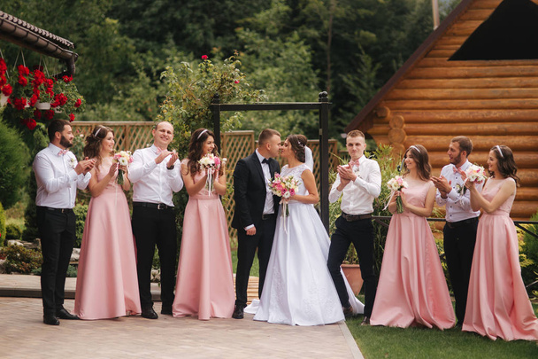 Groom and bride stand with groomsman and bridesmaid outside. Newlyweds kissing and friend clap. Wedding day - Photo, Image