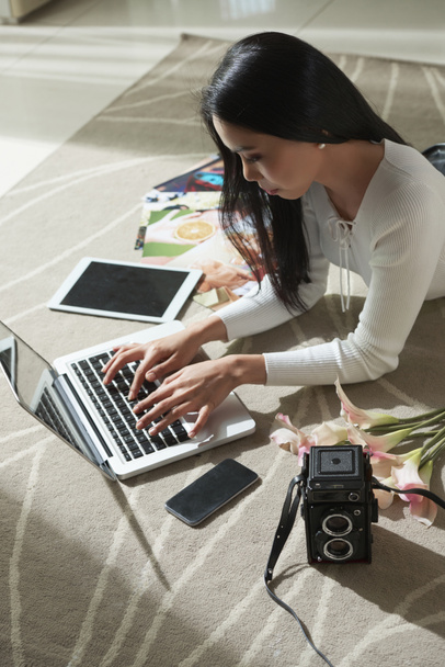 Νεαρή γυναίκα freelance φωτογράφος ξαπλωμένο στο πάτωμα στο στούντιό της και εργάζονται για το laptop - Φωτογραφία, εικόνα