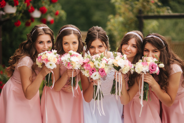 glückliche Braut mit Brautjungfer halten Blumensträuße und haben Spaß draußen. schöne Brautjungfer in gleichen Kleidern stehen der charmanten Braut im langen Hochzeitskleid zur Seite - Foto, Bild