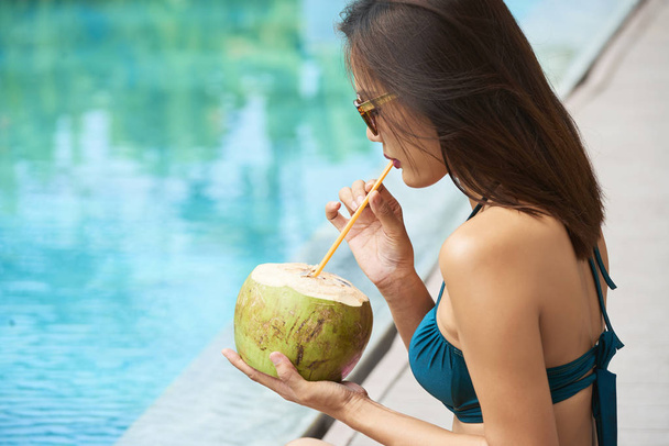 Young woman sitting at poolside and enjoying her drink outdoors during summer vacation  - Foto, afbeelding