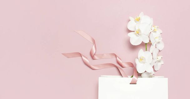 Flat lay top view Beautiful White Phalaenopsis orchid flowers and white gift bag on pastel pink background. Greeting card with orchid Tropical flower. Holiday birthday Womens Day floral Card beauty - Fotoğraf, Görsel