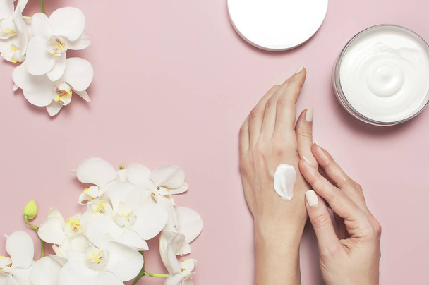 Young woman moisturizes her hand with cosmetic cream lotion opened container with cream body milk White Phalaenopsis orchid flowers on pink background Flat lay top view minimalism style Beauty concept - Photo, Image