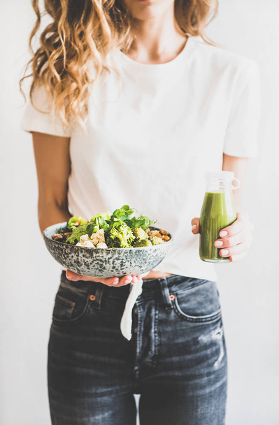 Healthy dinner, lunch. Woman in jeans standing and holding vegan superbowl or Buddha bowl with hummus, vegetable, fresh salad, beans, couscous and avocado and green smoothie in bottle - Φωτογραφία, εικόνα