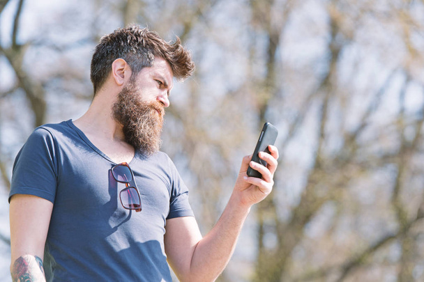 Hipster with beard using mobile phone outdoor. Man with beard and mustache on strict face calling, nature background, defocused. Bearded man typing message on cell phone. Communication concept - Fotoğraf, Görsel