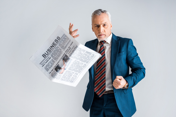 θυμωμένος ώριμος άνδρας σε επίσημη ένδυση ρίχνουν επιχειρηματική εφημερίδα ενώ στέκεται απομονωθεί σε γκρι  - Φωτογραφία, εικόνα