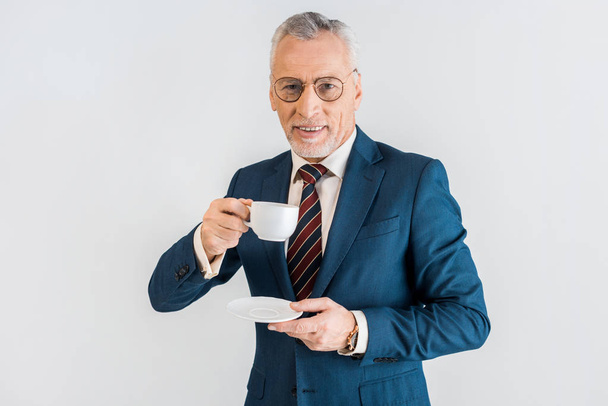 cheerful mature businessman in formal wear holding saucer and cup isolated on grey - Φωτογραφία, εικόνα
