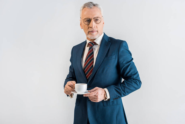 serious mature businessman in formal wear holding saucer and cup isolated on grey - Fotografie, Obrázek