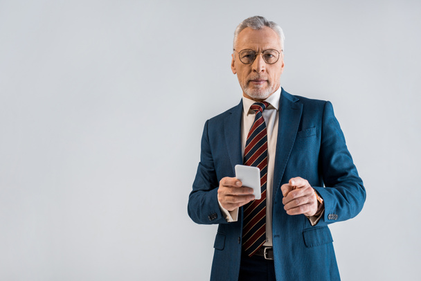 Reifer Geschäftsmann im Anzug hält Smartphone in der Hand und zeigt mit erhobenem Zeigefinger auf grau  - Foto, Bild