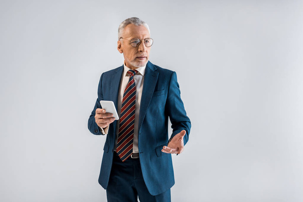 mature businessman in suit holding smartphone and gesturing isolated on grey  - Zdjęcie, obraz