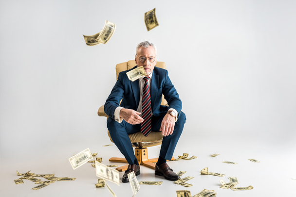 enfoque selectivo de hombre de negocios maduro sentado en sillón y lanzando billetes de banco en dólares de aire en gris
  - Foto, Imagen