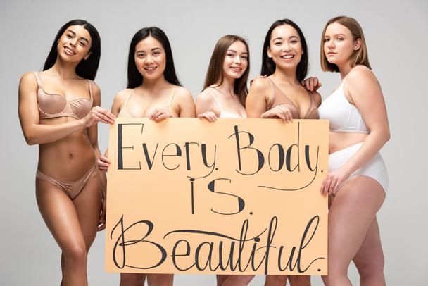 five smiling young multicultural women holding placard with "every body is beautiful" lettering isolated on grey - Photo, Image