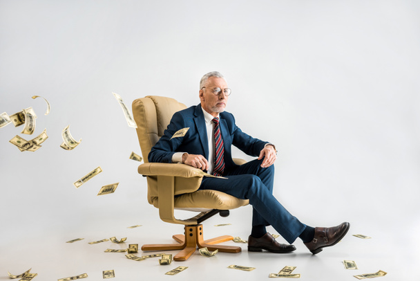 selective focus of confident mature businessman sitting in armchair near dollar banknotes on grey  - Zdjęcie, obraz