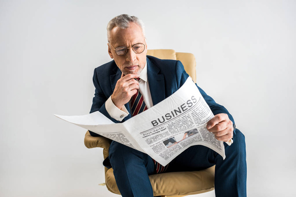 mature businessman sitting in armchair and reading business newspaper isolated on grey - Photo, Image