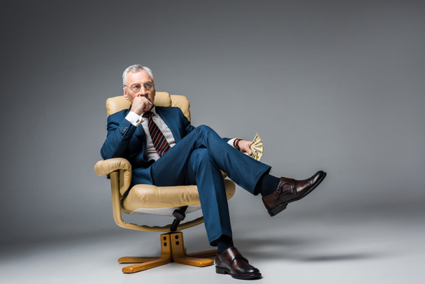 homme d'affaires mature réfléchi assis dans un fauteuil et tenant des billets en dollars sur gris
  - Photo, image