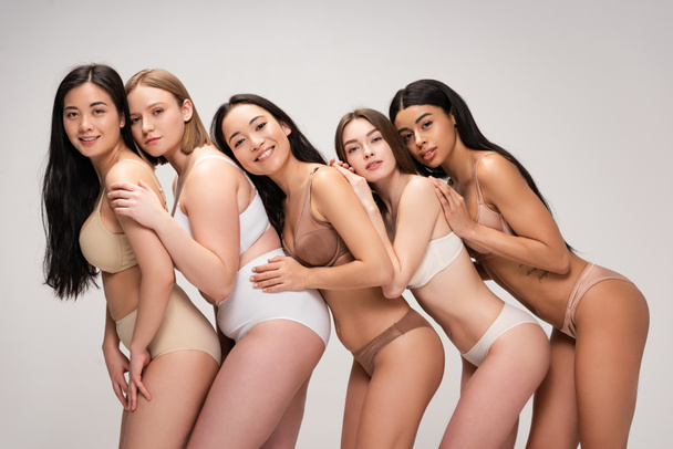 fünf glückliche junge multiethnische Frauen in Unterwäsche, die sich aneinander lehnen, isoliert auf grauem, körperpositivem Konzept - Foto, Bild