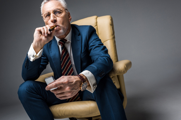Reifer Mann in Brille und Anzug mit Feuerzeug beim Zigarrenrauchen auf grau - Foto, Bild