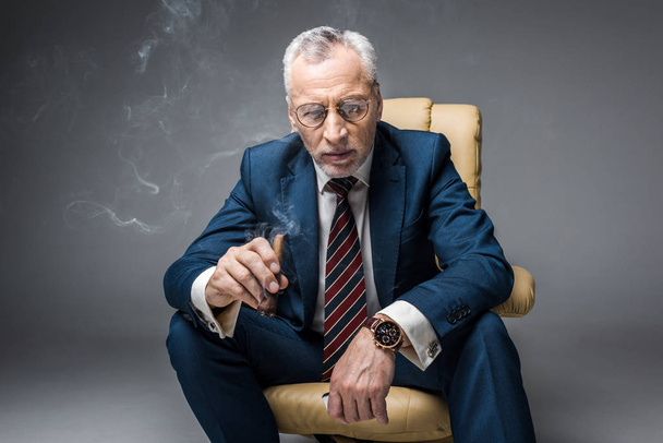 nachdenklicher reifer Geschäftsmann in Anzug und Brille mit Zigarre im Sessel auf grau - Foto, Bild