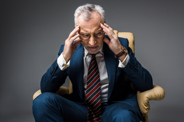 unavený zralé podnikatel v obleku s bolesti hlavy při sezení v křesle na grey  - Fotografie, Obrázek