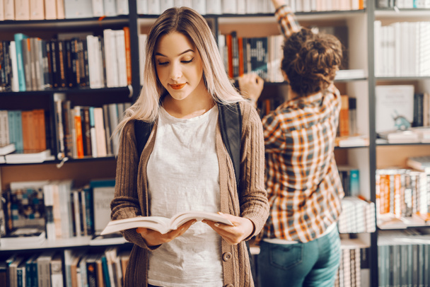 Pretty Caucasian girl standing in library with opened book and backpack. In background female student taking book from shelf. - Foto, Imagem