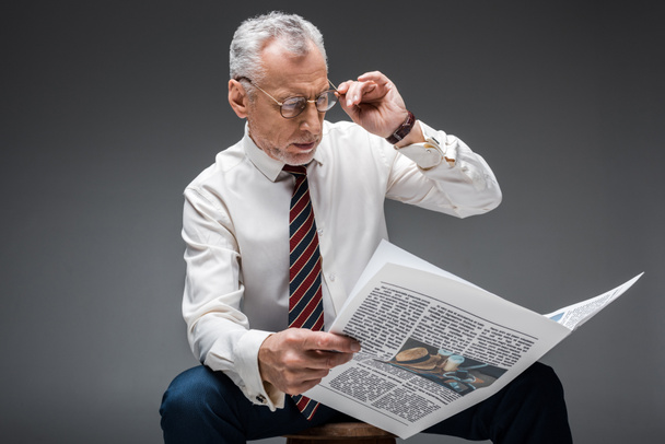 mature businessman touching glasses while reading newspaper on grey  - Фото, изображение