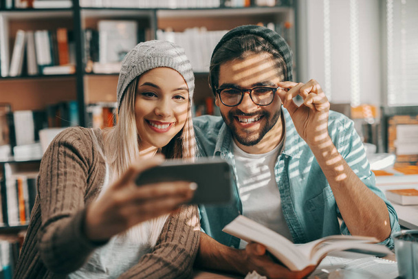 Két multikulturális csípő hallgatók selfie könyvtárban ülve pultnál. Ember tartja a könyvet, és a nő bevétel fénykép. - Fotó, kép