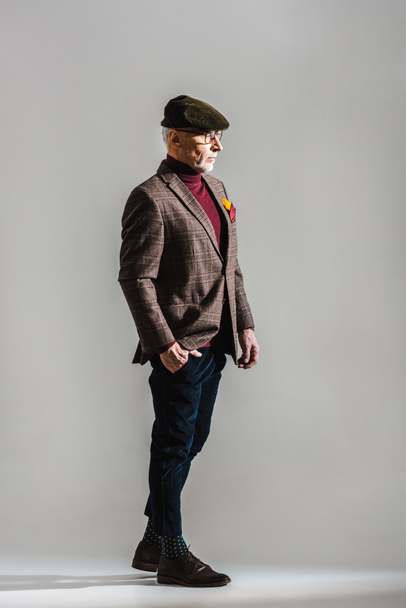 stylish middle aged man in glasses and cap standing with hand in pocket on grey  - Foto, Imagem