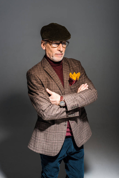 trendy middle aged man in glasses and cap standing with crossed arms on grey  - Foto, Imagen