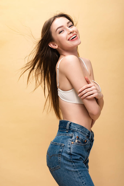 happy young woman in blue jeans and bra smiling and looking at camera isolated on beige - Фото, зображення