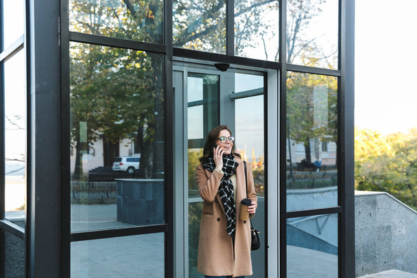 Woman outdoors walking by street drinking coffee talking by mobile phone. - Foto, imagen