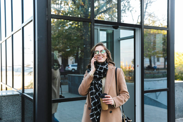 Woman outdoors walking by street drinking coffee talking by mobile phone. - Foto, Imagen