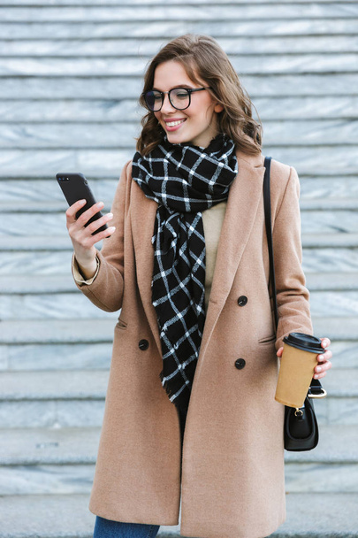 Feliz joven mujer al aire libre caminando por la calle bebiendo café usando el teléfono móvil
. - Foto, Imagen