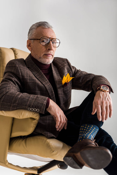enfoque selectivo de hombre maduro de moda en gafas sentadas en sillón con piernas cruzadas aisladas en gris
  - Foto, Imagen
