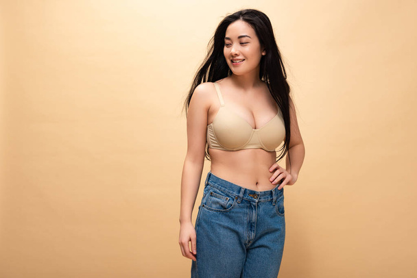 smiling young woman in blue jeans and bra holding hand on hip and looking away isolated on beige, body positivity concept - Φωτογραφία, εικόνα