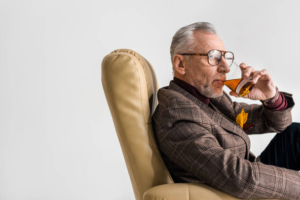 mature man sitting in armchair and drinking whiskey isolated on grey - Φωτογραφία, εικόνα