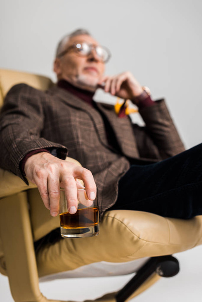 selective focus of glass with whiskey in hand of mature man in glasses sitting in armchair isolated on grey - Foto, Imagem